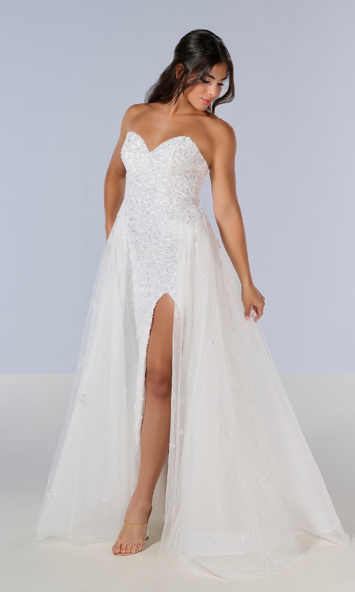 Wearing the Tiffany 16209 long prom dress, which features a strapless beaded bodice, sweetheart neckline, and high front slit, a woman stands against a plain background and looks down to her left.